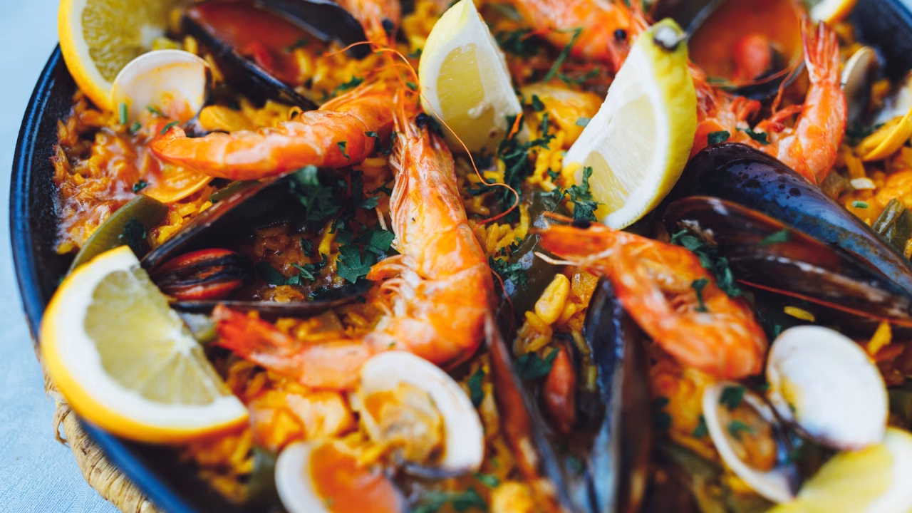 Dettaglio ravvicinato di una paella di pesce servita in padella, con gamberoni, cozze, vongole, riso allo zafferano, fette di limone e prezzemolo fresco. I colori vivaci degli ingredienti rendono il piatto invitante e tradizionalmente spagnolo.