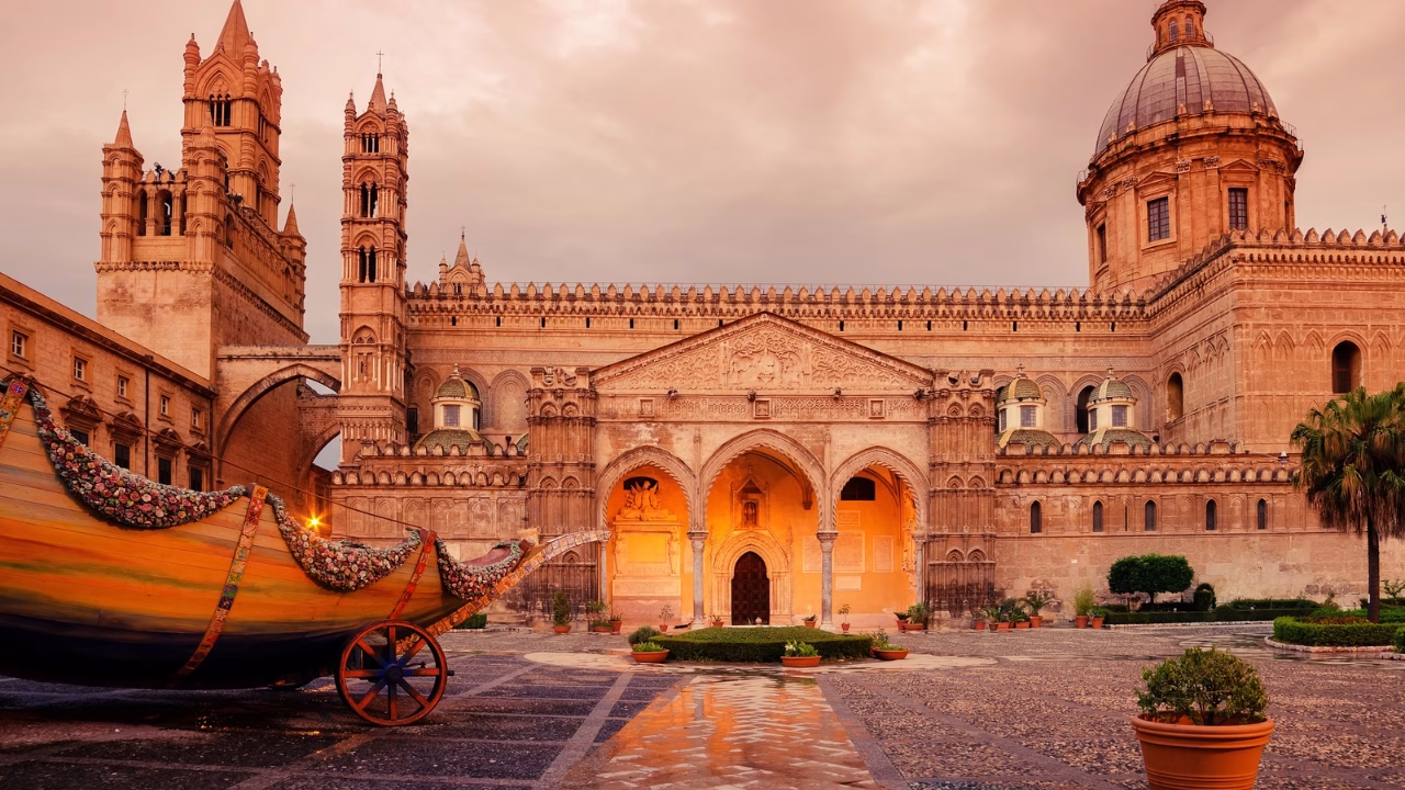 Cattedrale di Palermo all&rsquo;alba, illuminata da una calda luce arancione che esalta i dettagli architettonici arabi e normanni, con una barca decorata tipica siciliana in primo piano.