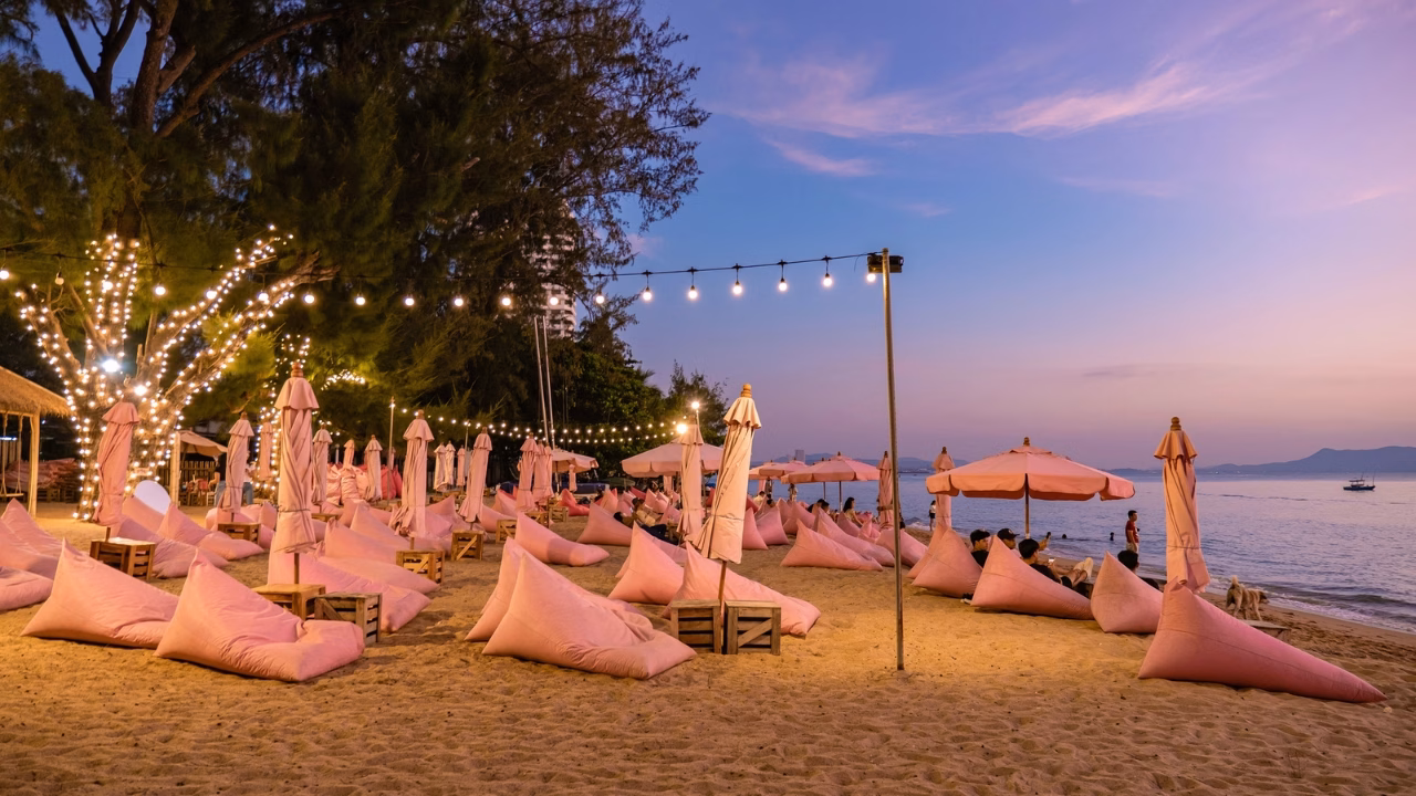 Lettini rosa e ombrelloni illuminati da luci soffuse sulla Dongtan Beach, la famosa spiaggia gay di Pattaya, al tramonto con il mare calmo sullo sfondo.