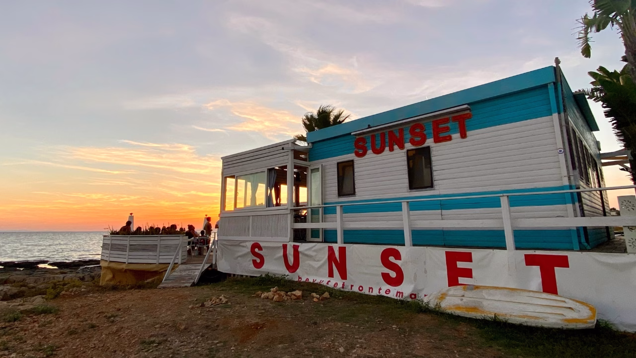 Chiosco bianco e azzurro con insegna rossa &ldquo;SUNSET&rdquo; affacciato sul mare, dove alcune persone si godono il tramonto sulle pedane esterne. Il cielo &egrave; tinto di arancio e rosa, creando un&rsquo;atmosfera perfetta per un aperitivo vista mare a Capilungo nel Salento.