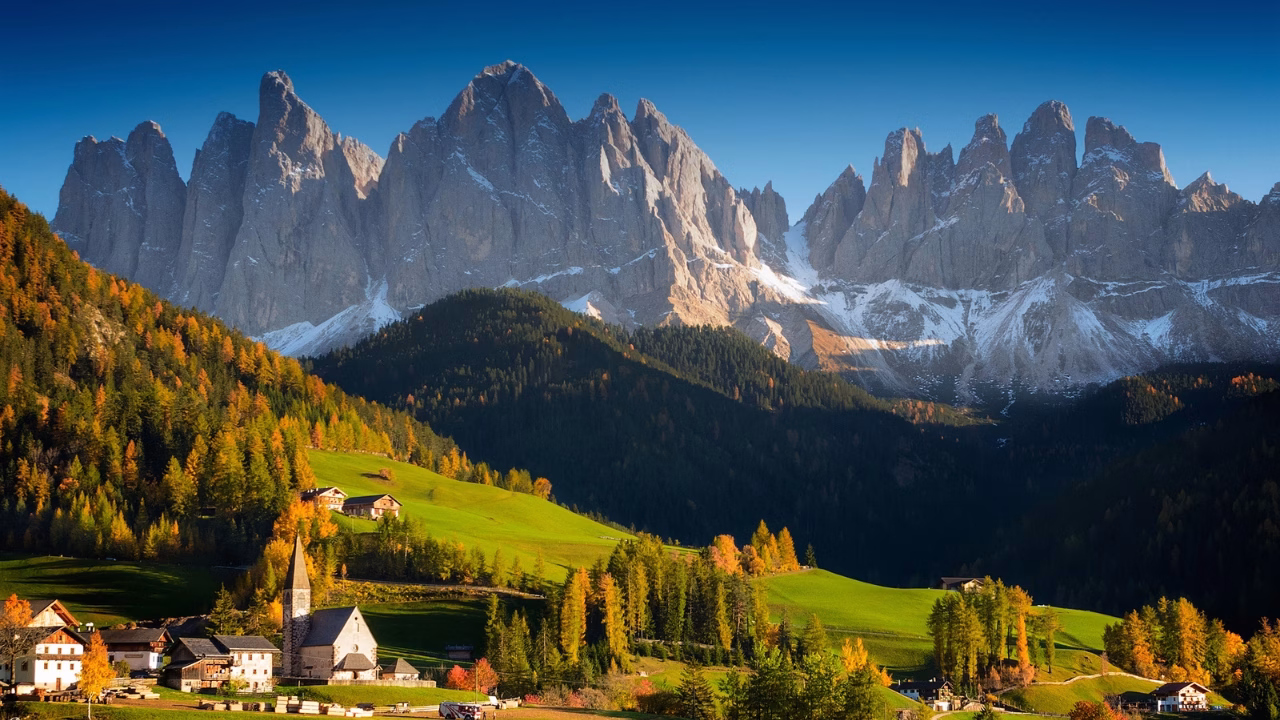 Veduta panoramica delle Dolomiti in autunno con le cime innevate sullo sfondo e un pittoresco villaggio alpino circondato da prati verdi e boschi dai colori caldi stagionali. Val di Funes.
