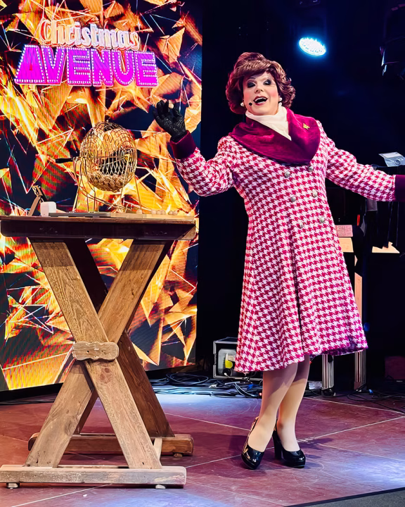 Drag queen conduce il Drag Bingo durante il mercatino di Natale gay Christmas Avenue a Berlino, indossando un abito a quadretti rosa e bianchi davanti a un tavolo da tombola.
