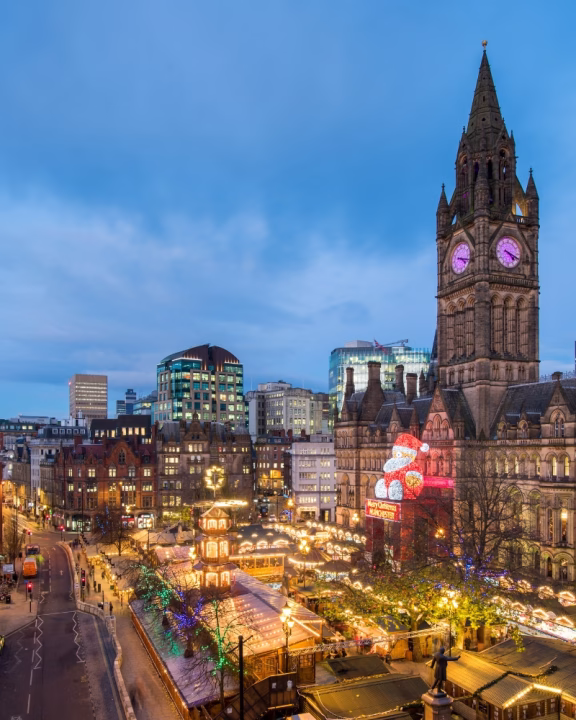 Vista panoramica dei mercatini di Natale LGBTQ+ di Manchester con il grande Babbo Natale arcobaleno illuminato sulla facciata del municipio e le luci festive che decorano gli stand nella piazza centrale.