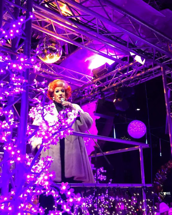 Drag queen avvolta in un cappotto bianco si esibisce su un palco illuminato da luci viola e decorazioni natalizie al mercatino LGBTQ+ Pink Christmas di Monaco di Baviera.