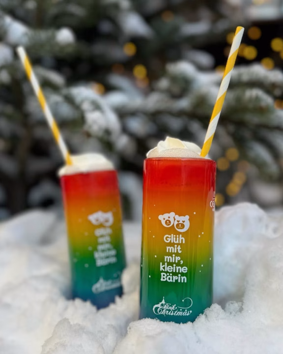 Due bicchieri arcobaleno con panna e cannucce colorate immersi nella neve, con la scritta &ldquo;Gl&uuml;h mit mir, kleine B&auml;rin&rdquo; e logo Pink Christmas, simbolo del mercatino di Natale LGBTQ+ di Monaco di Baviera.