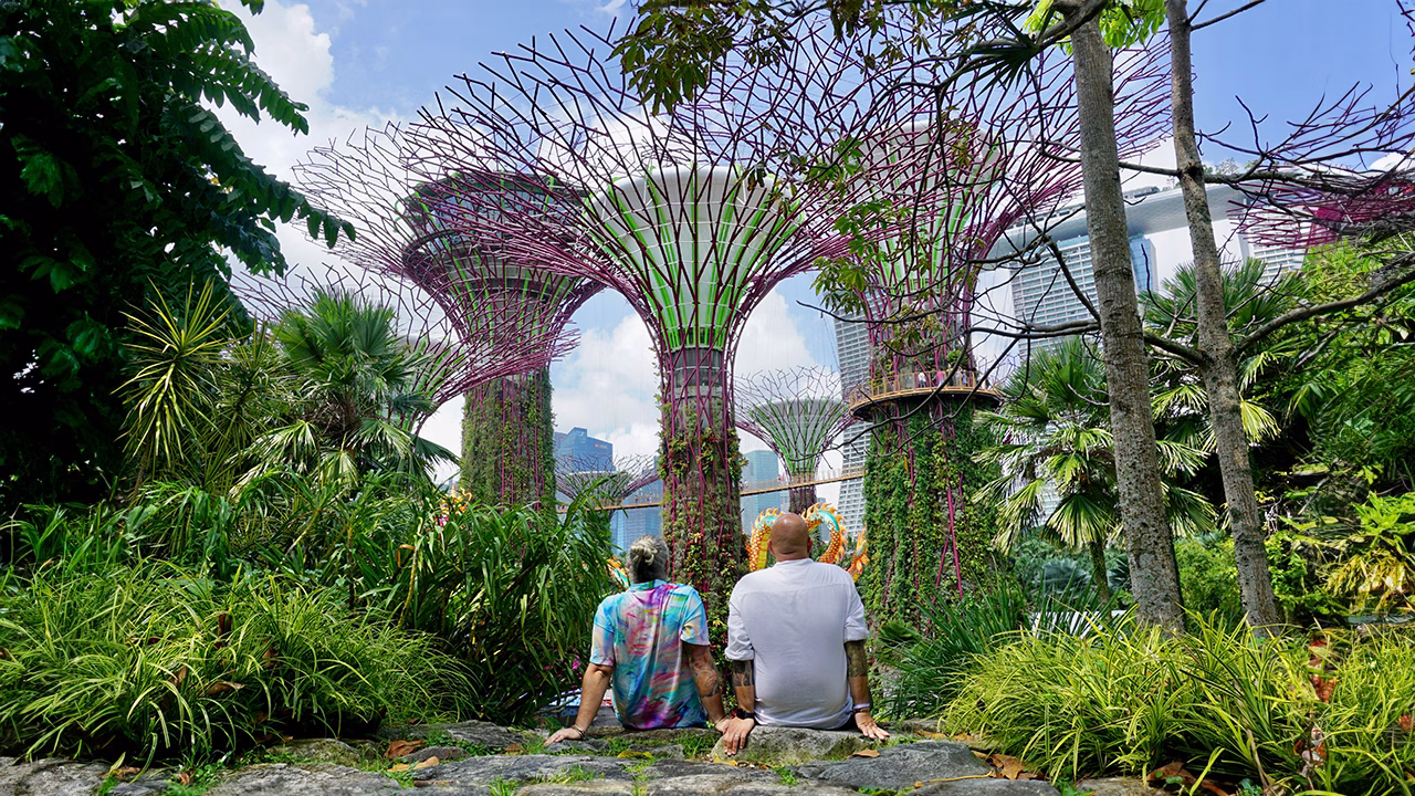 Daniele e Luigi di Gayly Planet, seduti nel verde rigoglioso del Gardens by the Bay di Singapore, ammirano i futuristici Supertree Grove in una scena da cartolina tropicale.