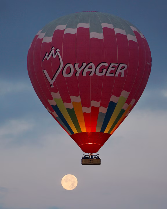 Mongolfiera colorata con la scritta &ldquo;Voyager&rdquo; che sorvola la Cappadocia all&rsquo;alba, con la luna piena visibile sullo sfondo del cielo.