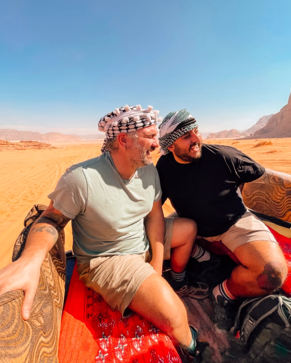 Daniele e Luigi di Gayly Planet sorridono seduti su un fuoristrada nel deserto del Wadi Rum, in Giordania, con copricapo tradizionali e paesaggio arido alle spalle