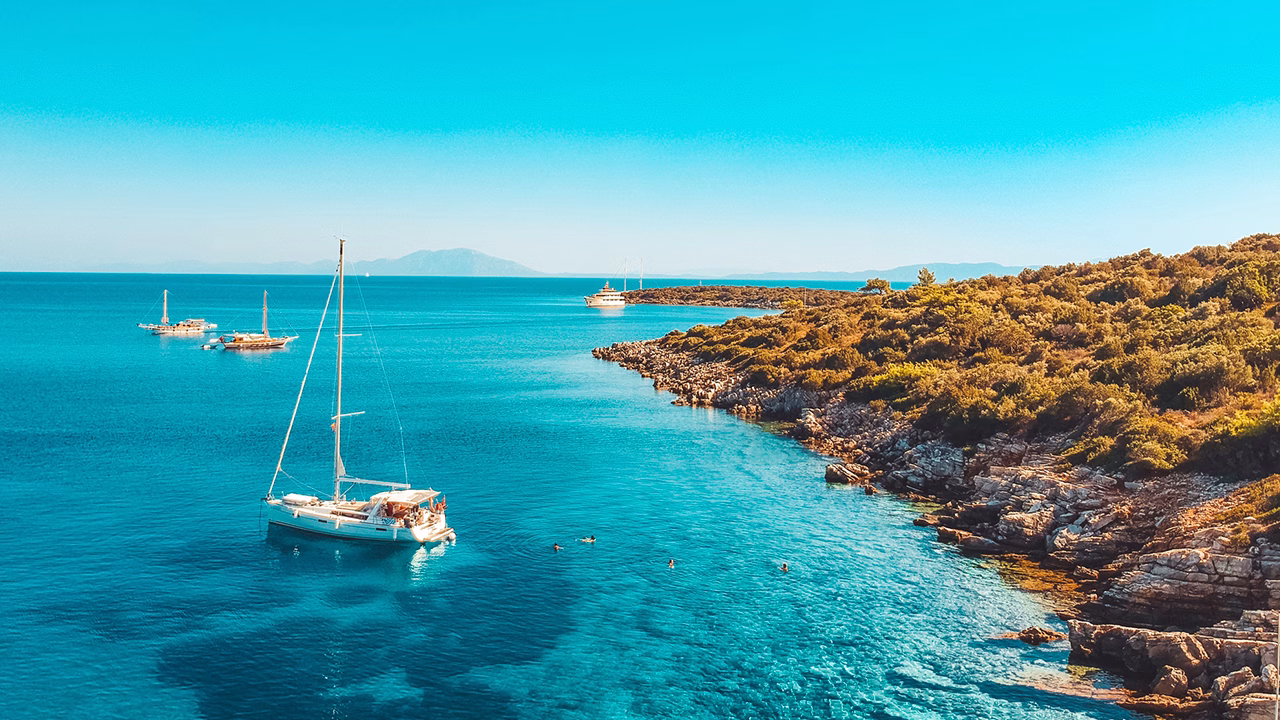 Barche a vela e yacht ormeggiati nelle acque cristalline di Bodrum, lungo una costa rocciosa punteggiata da vegetazione mediterranea, sotto un cielo terso e soleggiato.