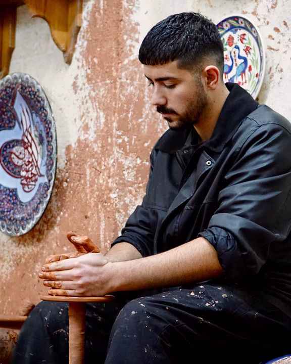 Giovane artigiano turco lavora l&rsquo;argilla con le mani su un tornio in una bottega decorata con piatti in ceramica dipinti a mano, durante una dimostrazione a G&ouml;reme