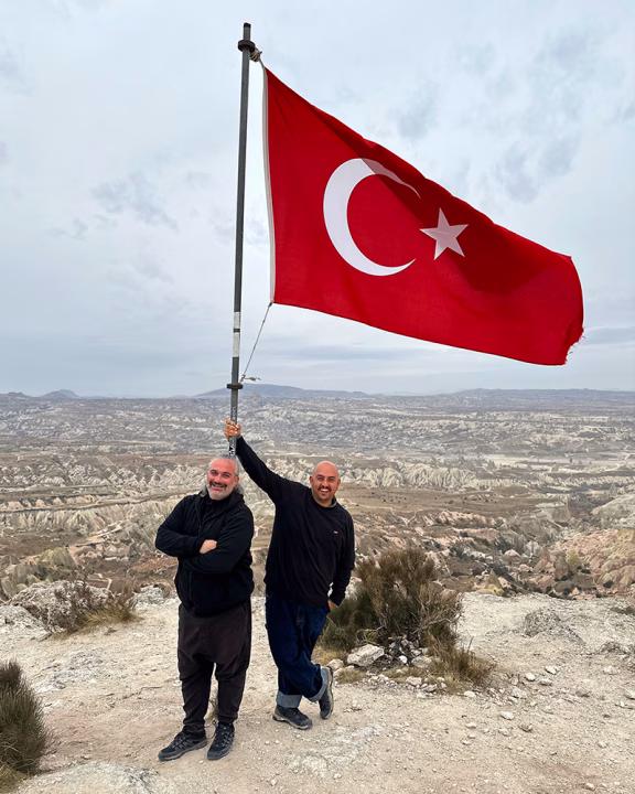 Daniele e Luigi di Gayly Planet sorridenti posano accanto a una grande bandiera turca che sventola su un&rsquo;altura, con il paesaggio roccioso e lunare della Cappadocia sullo sfondo