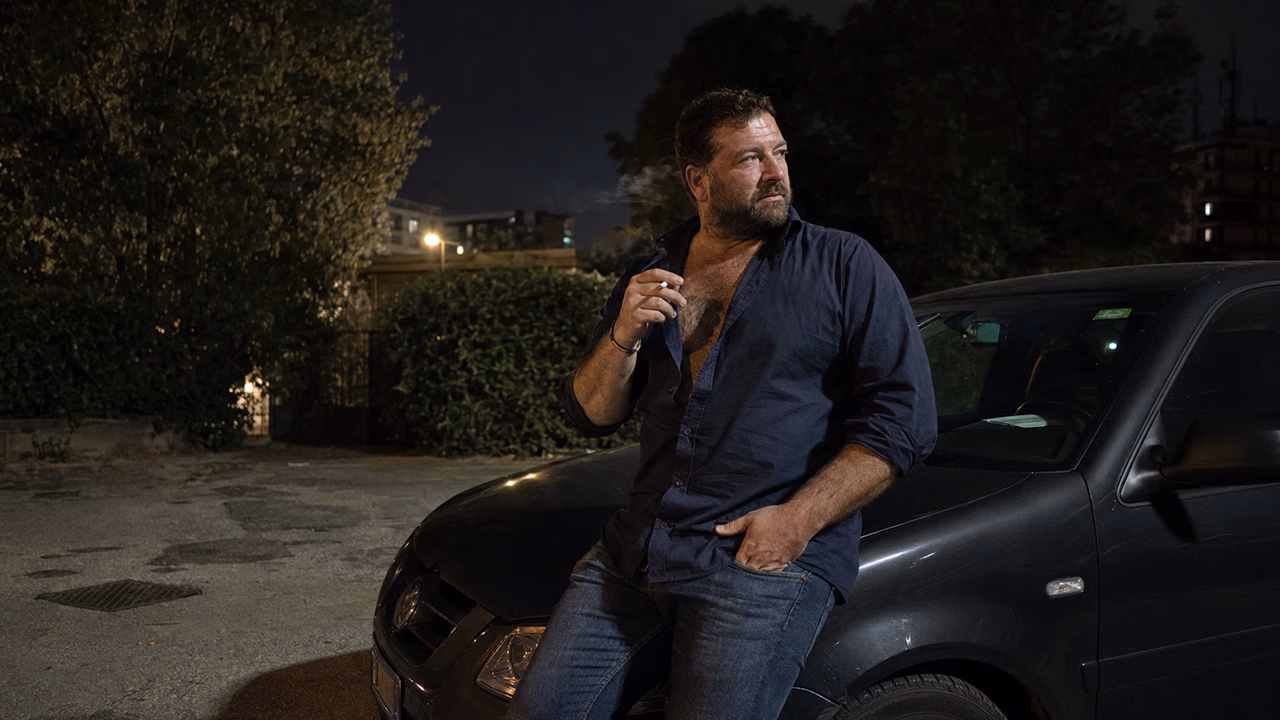 Uomo gay adulto stile bear fuma appoggiato a un’auto in un parcheggio isolato di città - Immagine a scopo illustrativo generata con IA